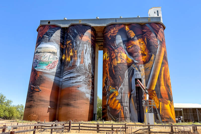 Four large grain silos feature vibrant Silo Art murals, including a man in a hat holding tools and a striking landscape scene, all set beneath a clear blue sky.
