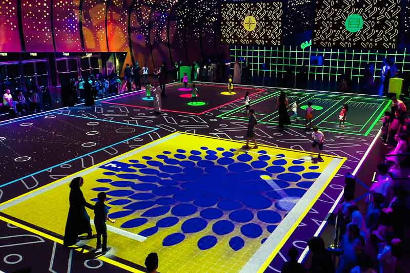 People walk and play on a large, interactive, neon-lit sports court with colorful digital projections and geometric patterns on the floor and walls.
