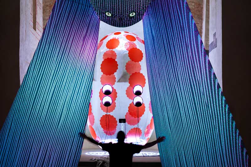 A person stands with arms outstretched in front of a large, illuminated abstract installation with red circles and blue hanging strands.