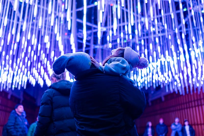 A person holding a child in winter clothing stands under a ceiling of hanging blue LED lights, with several people visible in the background.