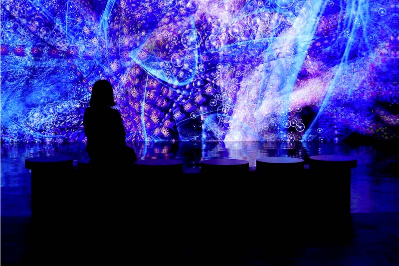 A person sits in silhouette on a bench, facing a bright, abstract blue and purple digital art display filled with swirling patterns and light effects.