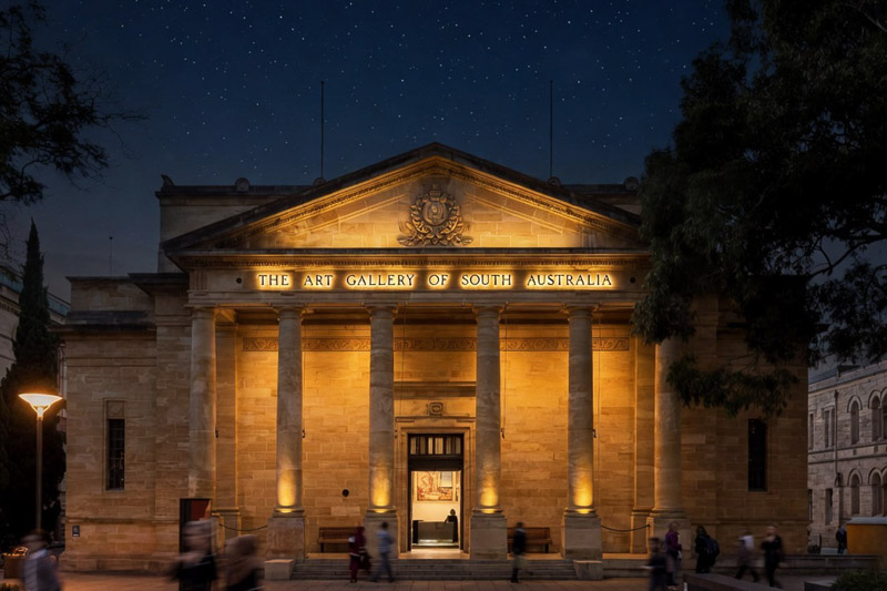 The Art Gallery of South Australia is illuminated at night, showcasing its neoclassical facade with columns and a lit entrance under a starry sky.