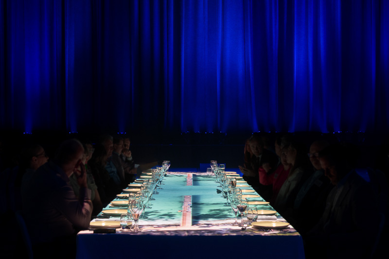A long dining table set with plates, glasses, and cutlery, surrounded by seated people, under dramatic blue stage lighting.