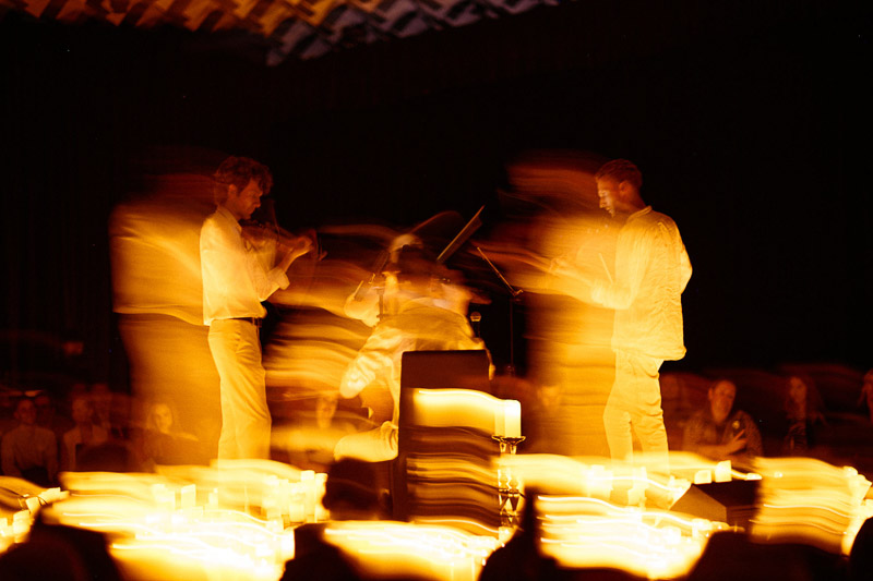 A string quartet performs surrounded by blurred, glowing candlelight, with motion blur creating a dynamic effect; audience members watch in the background.