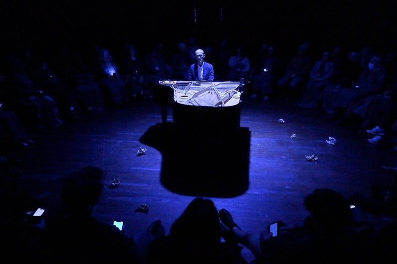 A person plays a grand piano under a blue spotlight, surrounded by an audience seated in a dark room.